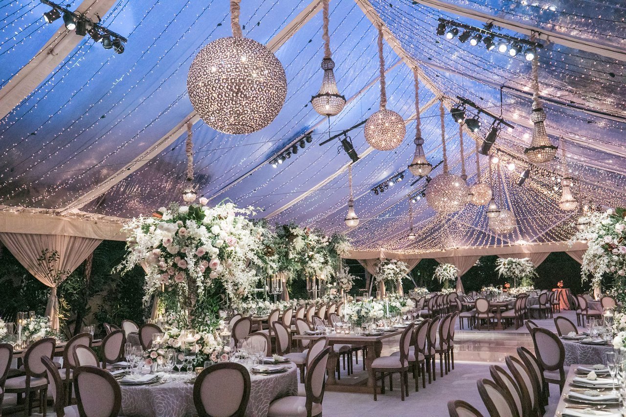 A wedding reception beneath a clear tent filled with chandeliers and twinkling lights